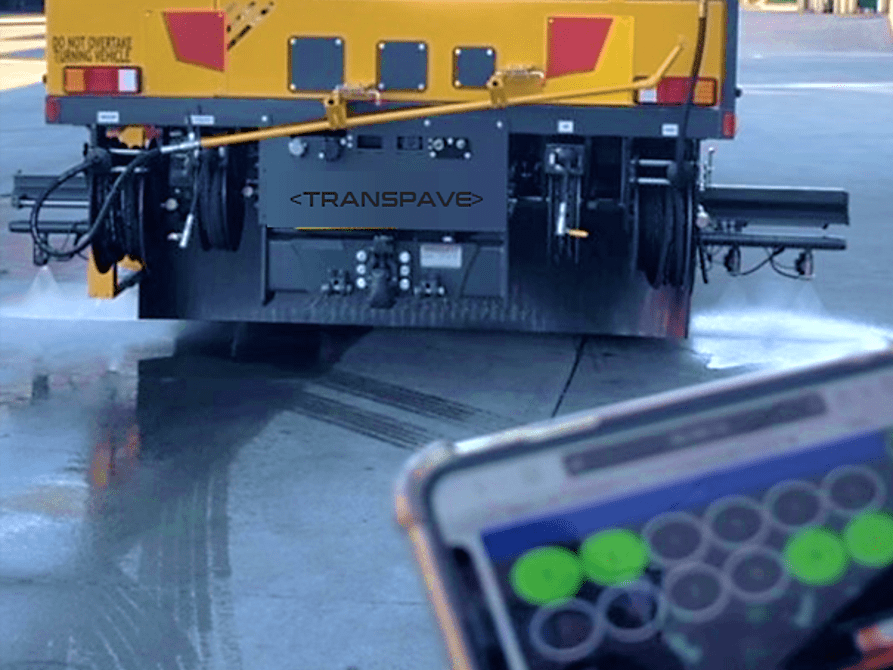 Transcoater Tack Coater spraying material from extended spray bar in the background, mobile phone controlling spray bar in the foreground